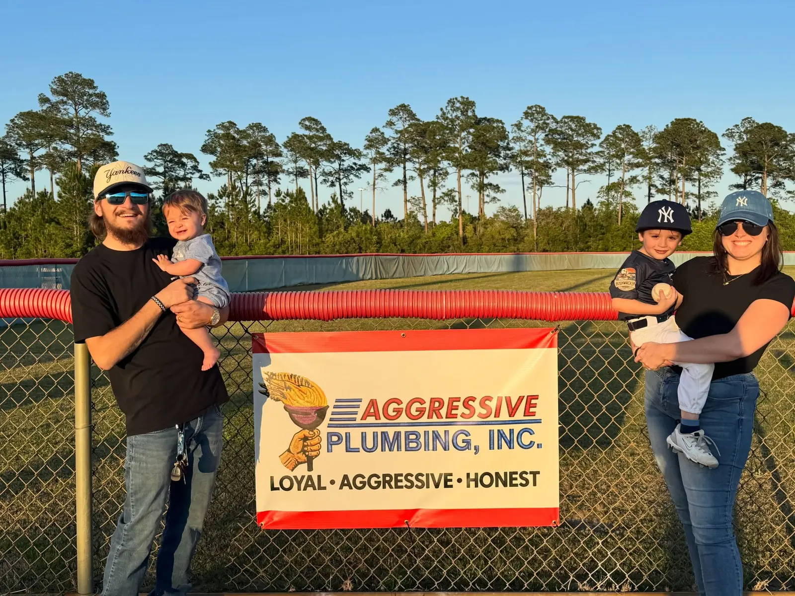 Local families standing by the Aggressive Plumbing sponsor banner at a Pensacola ballfield